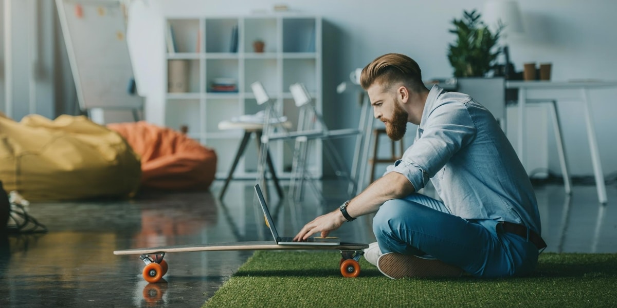 A man using a laptop perched on a skateboard in a light airy apartment. It looks like he is apply for a new job in Summer