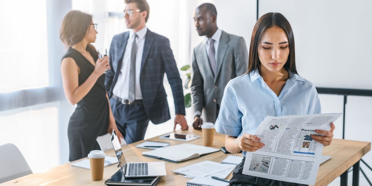 a woman looks at the newspaper while her colleagues at the back chat about the tech industry