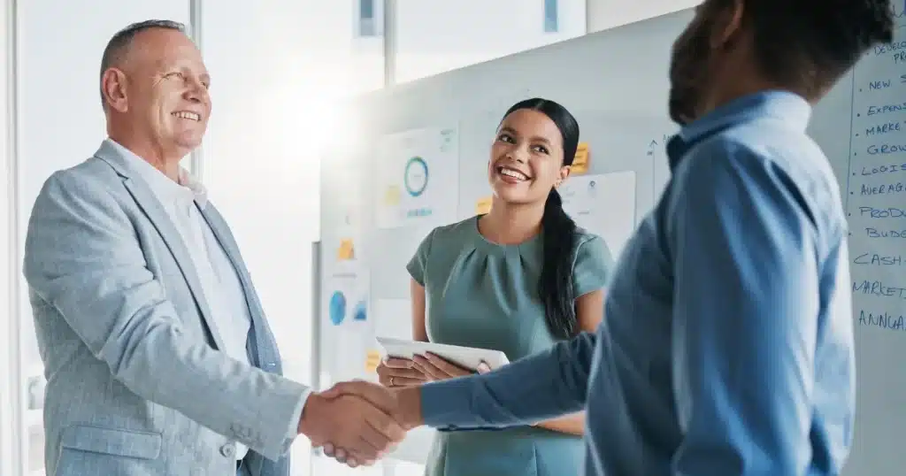 A white man and a black man shake hands and a woman smiles in the background. It's an office environment, after a business agreement.