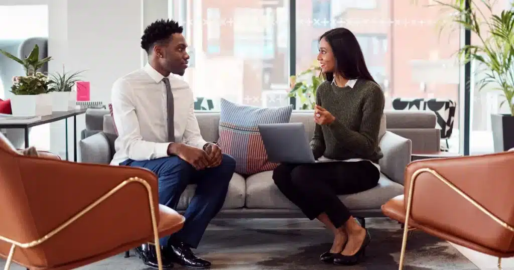 Two professionals sat and a sofa. The woman holds a laptop and is performing an informal exit interview to a male colleague who has resigned.