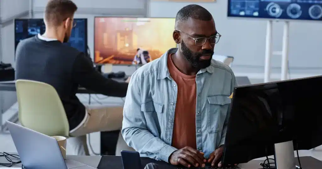 A black game developer codes in his computer while a game designer tests the game on the background