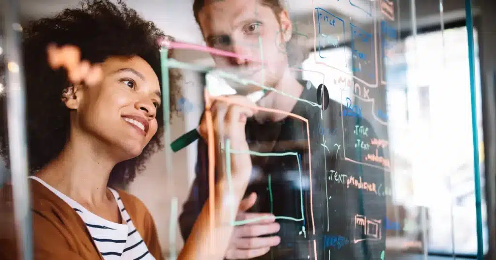 A racialised woman writes takes notes on a web design on a board while her male colleague offers his feedback