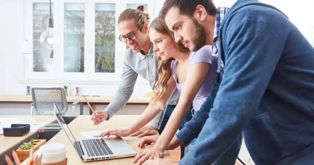 Two young lads and a young woman lean over the table with a laptop, learning together and taking notes