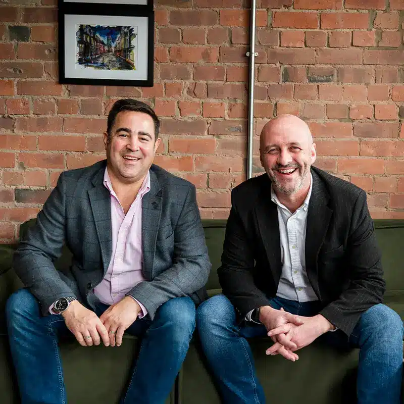 Nick Derham and David Berwick, Adria Solutions Directors, smile confident at the camera, sat on a sofa